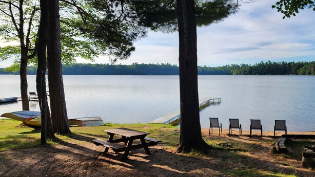A lakeside picnic area with a table and chairs, surrounded by trees and a view of the lake. - Reggie's Northwoods Retreat (St Germain)