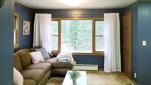 A living room with a brown sectional sofa, a small table with a plant, and a window with curtains. - Reggie's Northwoods Retreat (St Germain)