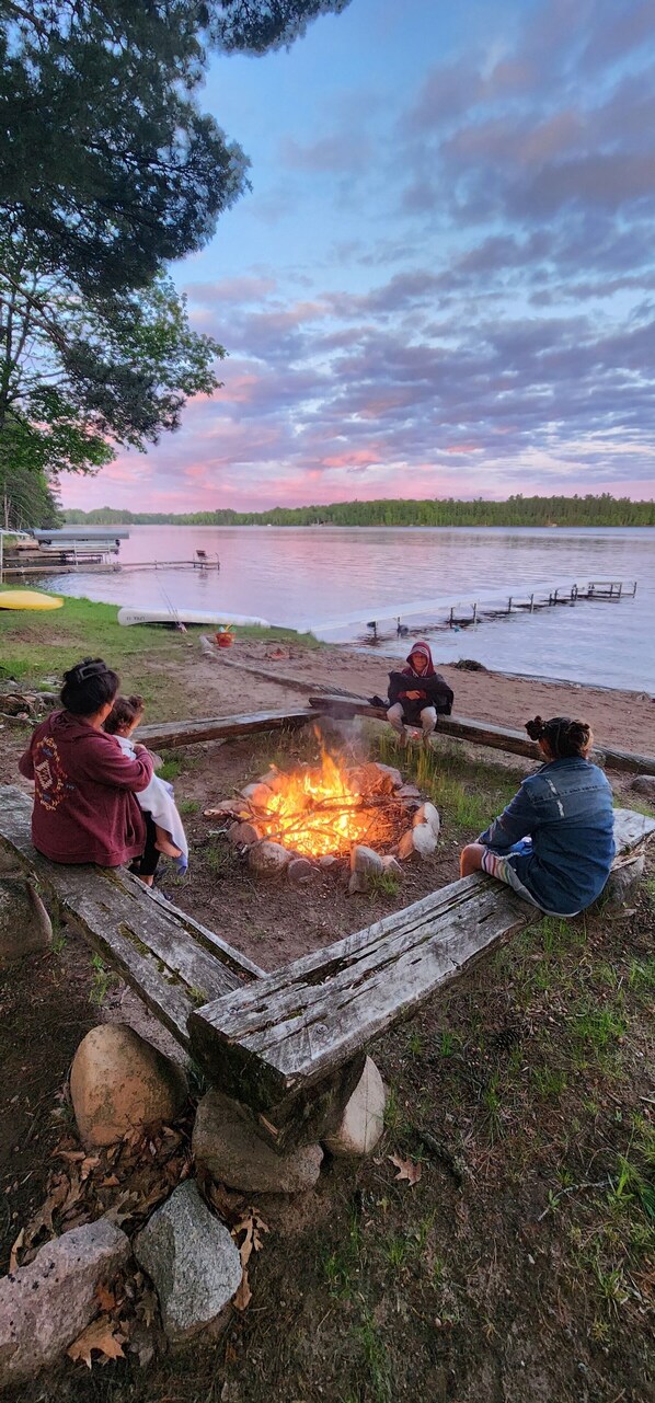 A campfire with people sitting around it, overlooking a lake with boats and docks in the background. - Reggie's Northwoods Retreat (St Germain)