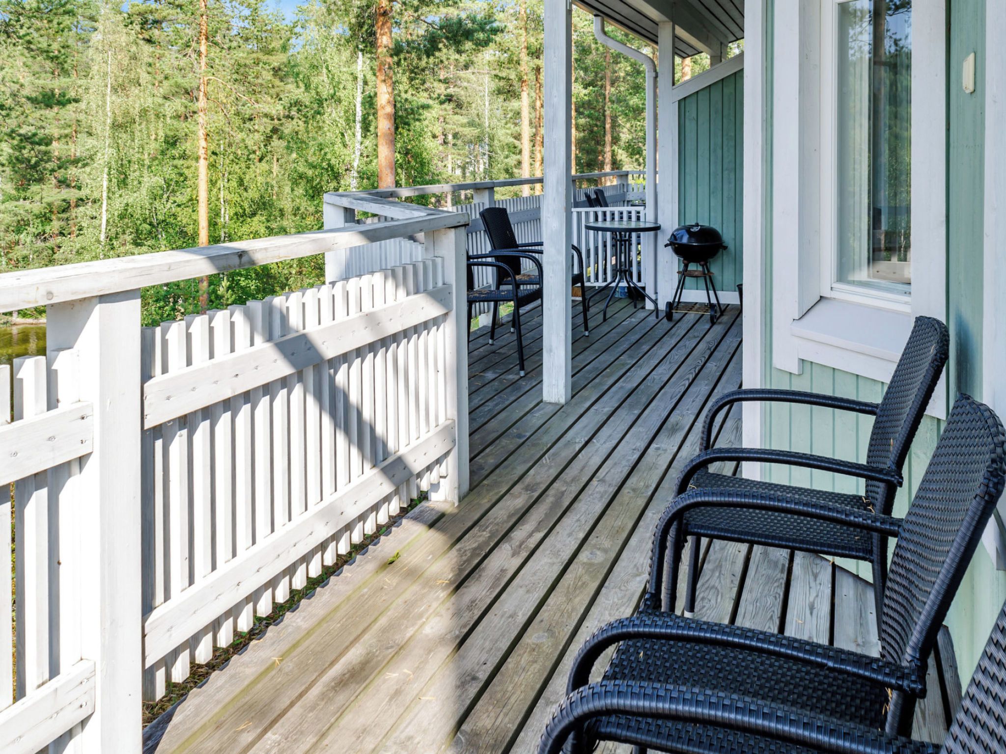 Eine Holzterrasse mit weißer Brüstung, Gartenmöbeln und Blick auf eine bewaldete Gegend.