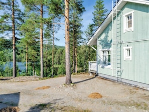 Een huis met een grindpad, een houten terras en uitzicht op een meer, omringd door hoge dennenbomen.