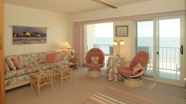 A living room with a sofa, two armchairs, a coffee table, and a side table. There is a painting on the wall and a window with a view of the sea.