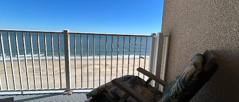 A balcony with a view of the beach, a chair, and a textured wall.