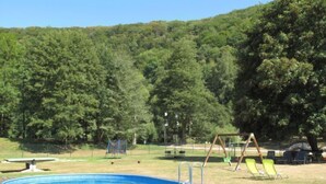 An outdoor swimming pool with a wooden deck, surrounded by a grassy area with trees and playground equipment. - Green Valley Park Stárkov by Interhome (Stárkov)