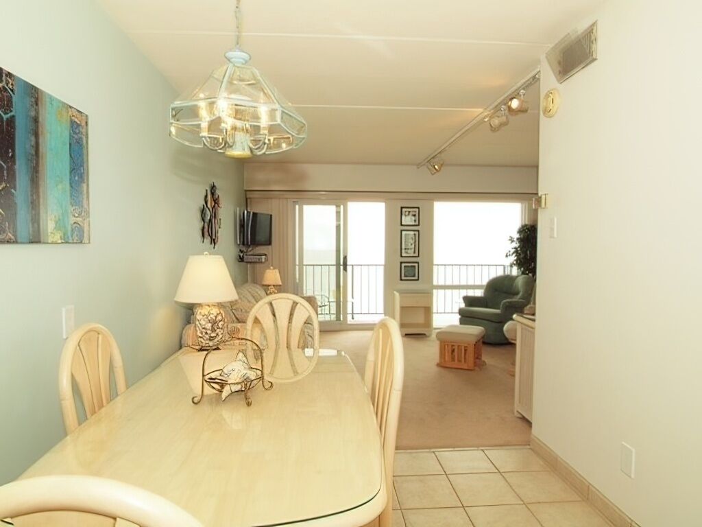 A dining area with a table, chairs, and a chandelier. There is a living room in the background with a sofa and a television.