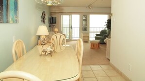 A dining area with a table, chairs, and a chandelier. There is a living room in the background with a sofa and a television.