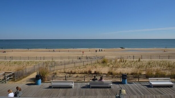 Uitzicht op het strand vanaf een houten terras met banken en een hek, onder een heldere lucht.