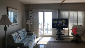 Een woonkamer met een bank, een televisie, een lamp en een balkon met schuifdeuren.