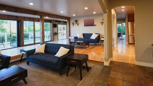 A spacious living area with a dark blue sofa, a coffee table, and a dining table. There are large windows providing a view of the outdoors. - No Cleaning Fee! Luxury Broadmoor Home w/ Hot Tub & Fire Pit (Broadmoor)
