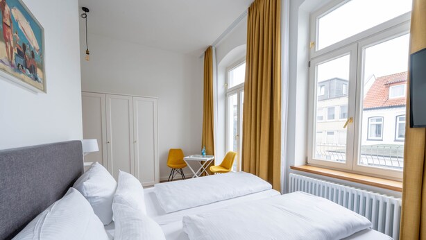 A hotel room with a large bed, a small table with two chairs, and a window with curtains. - Double room - Logierhaus am Rathaus (Norderney)