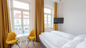 A hotel room with a bed, two yellow chairs, a TV, and a window with curtains. - Double room - Logierhaus am Rathaus (Norderney)