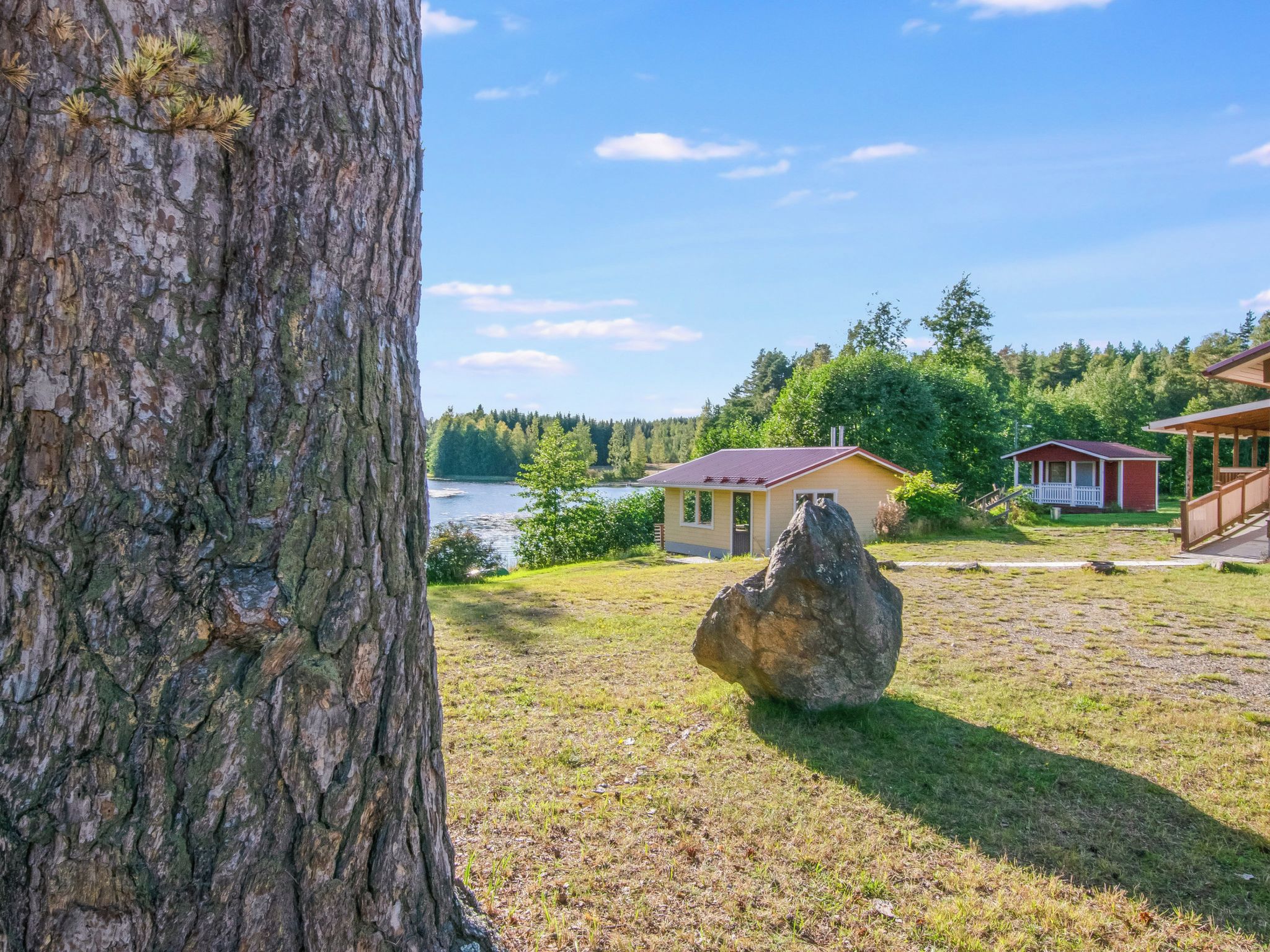 Järvenrantamökki, jonka edessä on suuri kivi, ja jonka ympärillä on nurmikkoalue ja taustalla metsä.