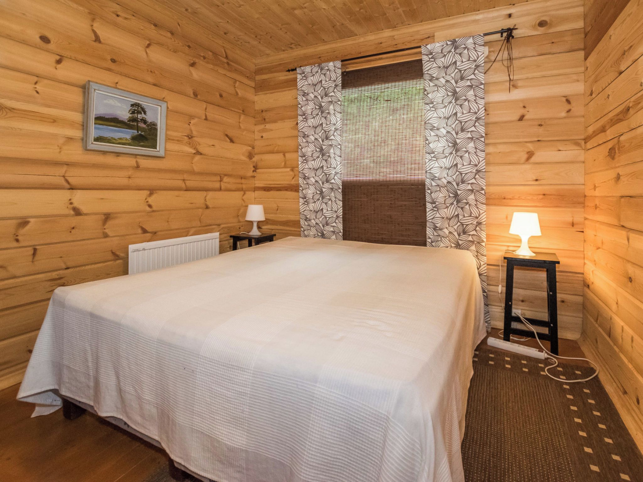 A wooden cabin room with a bed, two bedside tables with lamps, a radiator, and a window with patterned curtains.