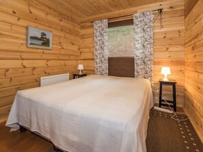 A wooden cabin room with a bed, two bedside tables with lamps, a radiator, and a window with patterned curtains. - Kuusela by Interhome (Somero)