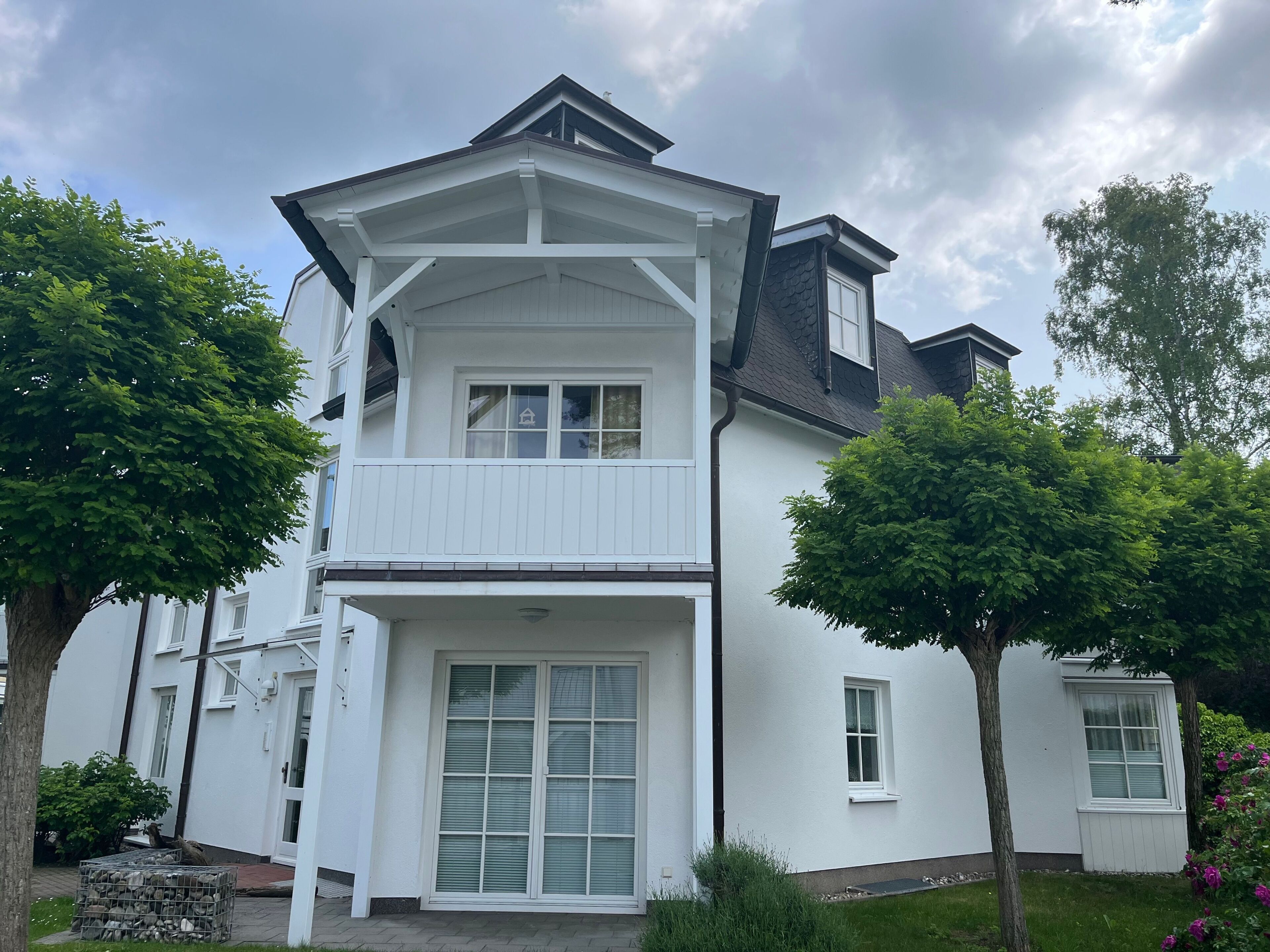 Ein weißes Haus mit zwei Etagen, einem Balkon, einer großen Haustür und einem kleinen Fenster im oberen Stockwerk.