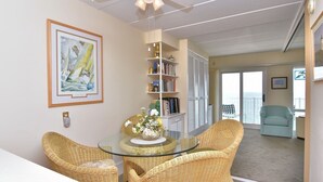 Ein Wohnzimmer mit Rattanmöbeln, einem Glastisch, einem Deckenventilator, einem Bücherregal und Blick ins Freie.