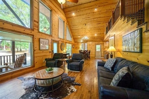 A spacious log cabin interior with a large dining table, multiple seating areas, and large windows offering views of the outdoors. - Luxury 2BD Cabin w/ Hot Tub & Game Loft (Broken Bow)