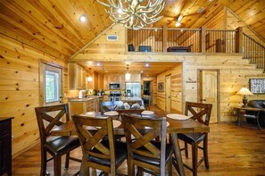 A wooden cabin interior with a dining area, kitchen, and upper balcony.