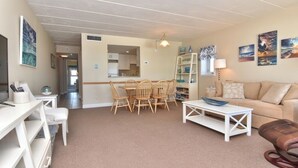 A living room with a beige sofa, a coffee table, a dining table with chairs, a white TV stand, and a bookshelf.