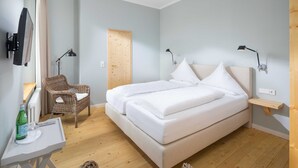 A bedroom with a bed, two bedside tables, a chair, a desk, a television, and a door. - Comfort Double Room - Logierhaus Oystercatcher (Norderney)