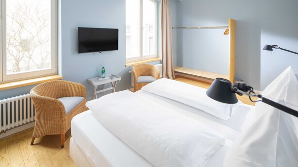 A hotel room with a bed, a TV, a chair, a small table, and a window with curtains. - Comfort Double Room - Logierhaus Oystercatcher (Norderney)
