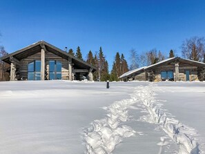 Uma cabana de troncos com uma grande porta de vidro e uma chaminé de pedra, rodeada por neve e árvores.