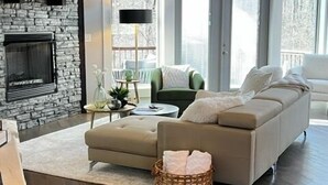 A spacious living room with a stone fireplace, a green sofa, a beige couch, and a large window with a view of trees.