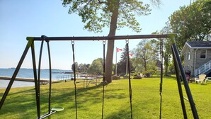 A swing set in a grassy area with a view of a body of water and a building in the background. - Home of 1000island#3-Directly on STL river private beach FREE boat docking (Gananoque)