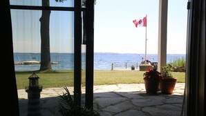 A view from a window overlooking a lake with a dock, a Canadian flag, and potted plants on a stone patio. - Home of 1000island#3-Directly on STL river private beach FREE boat docking (Gananoque)