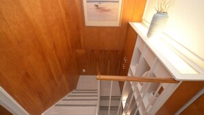 Una escalera de madera con barandilla, un cuadro enmarcado en la pared y un florero con ramas en el alféizar de una ventana.