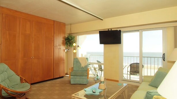 Una sala de estar con sofá, una butaca, una mesa de centro y televisor. Se ve el océano a través de grandes ventanales.