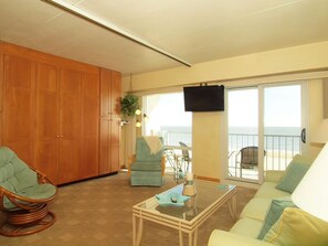 A living room with a sofa, a chair, a coffee table, and a television. There is a view of the ocean through large windows.