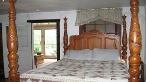 Uma cama com dossel, com colcha estampada com motivos florais, cabeceira de madeira e baldaqueim rendado sobre a janela.