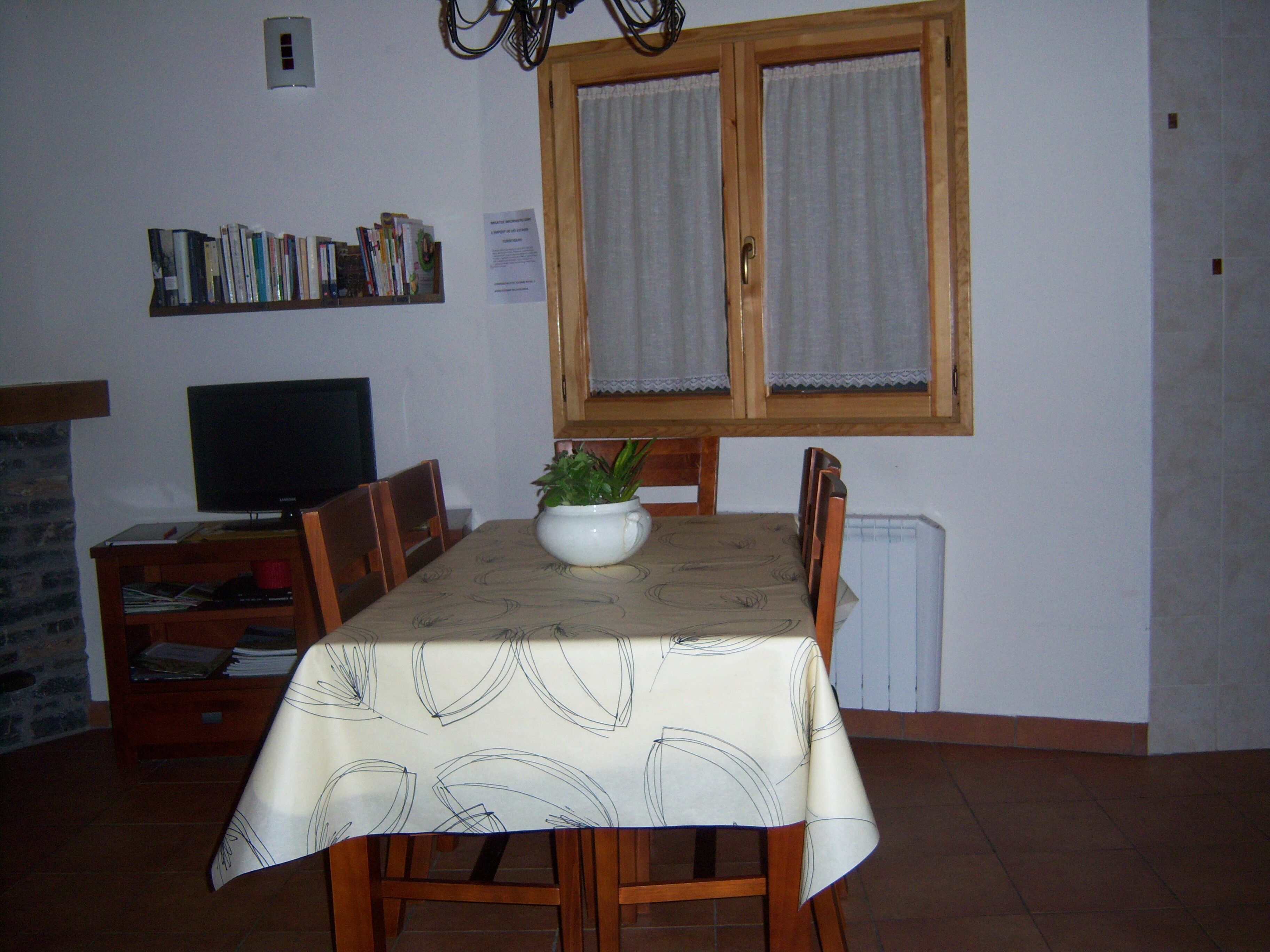 A dining area with a table set for four, a television on a stand, a bookshelf with books, and a window with curtains.