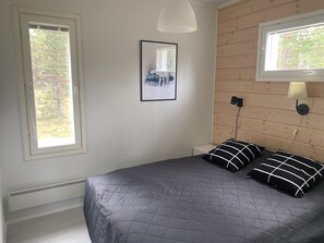 Un dormitorio con cama, dos lámparas de noche, un cuadro enmarcado en la pared y ventanas con vistas a árboles.