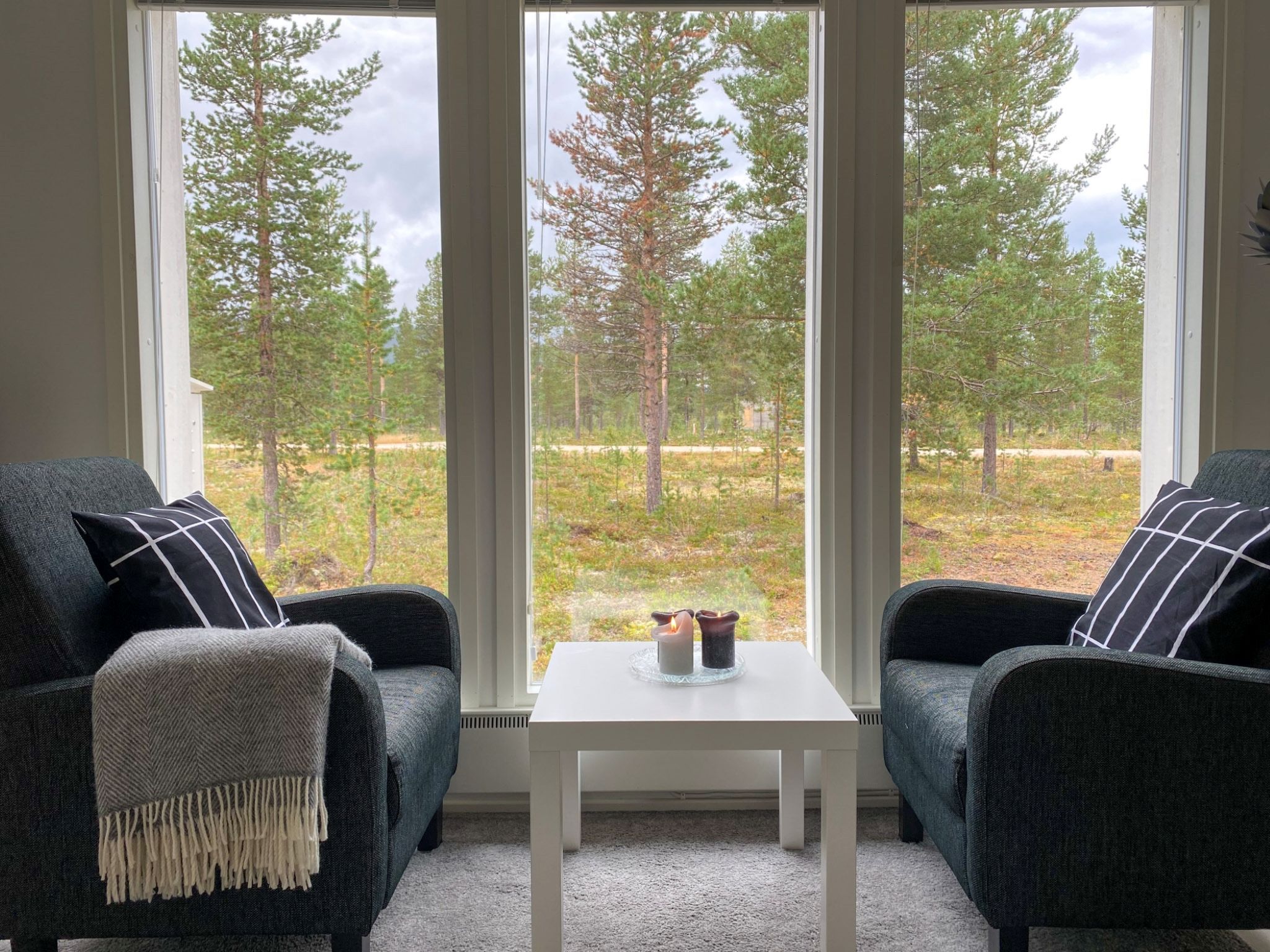 A room with two dark armchairs facing a white table with candles, large windows offering a view of a forest.