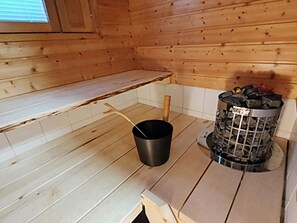 Interior de uma cabana de madeira, com um balde de metal, uma colher de madeira e uma lixeira com a etiqueta 'Hervia'.