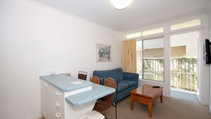A small, well-lit room with a kitchenette, a sofa, a coffee table, and a large window offering a view of a balcony. - Beachside Shoal Bay Unit 4 (Shoal Bay)