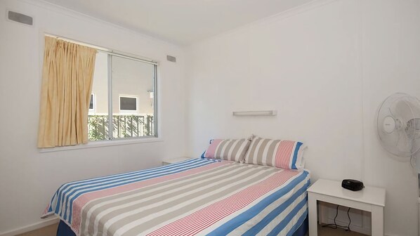 Un dormitorio con cama, ventana con cortinas, mesita de noche con ventilador y vistas a un balcón.
