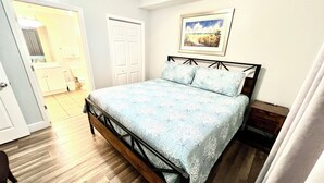 Un dormitorio con cama, piso de madera, puerta blanca, baño y un cuadro enmarcado en la pared.