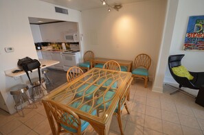 A compact kitchen with modern appliances, a dining area with a glass-top table and wicker chairs, and a black chair with a green cushion.