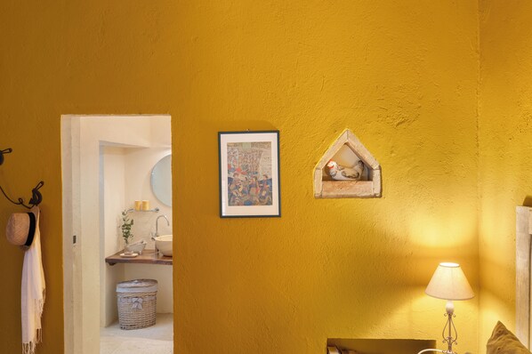A bathroom with a yellow wall, a framed picture, a small decorative house, a lamp, and a towel hanging on the wall.