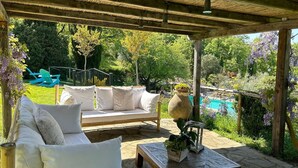 Un espace extérieur aménagé, abrité et doté d’un banc en bois et d’un canapé blanc, entouré d’une végétation luxuriante et d’une piscine.