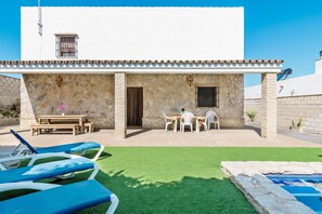 A building with a stone facade, a patio with a table and chairs, and a pool area with lounge chairs.