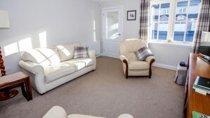 A living room with a white sofa, a cream armchair, a wooden side table, and a window with a view of houses. - Devonvale Place by Interhome (Kinross)