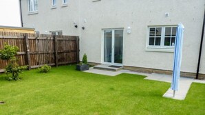 A residential backyard with a wooden fence, a small garden, and a patio area with a blue-striped umbrella. - Devonvale Place by Interhome (Kinross)