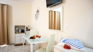 Una habitación pequeña y limpia con una mesa de comedor, un sofá, un televisor montado en la pared y una zona de cocina con cafetera y fruta.