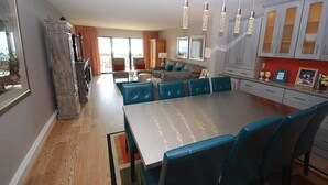 A spacious dining area with a long table and blue chairs, a kitchen with white cabinets, and a living area with a sofa and a TV. - Edgewater House - Ocean Front Units With Spectacular Views! (Rehoboth Beach)