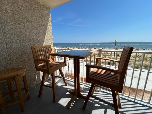 A balcony with two wooden chairs and a table, overlooking a beach with umbrellas and people.