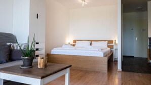 Moderni hotellihuone, jossa on puinen sänky, valkoiset seinät ja pieni pöytä, jonka päällä on kasvi.
